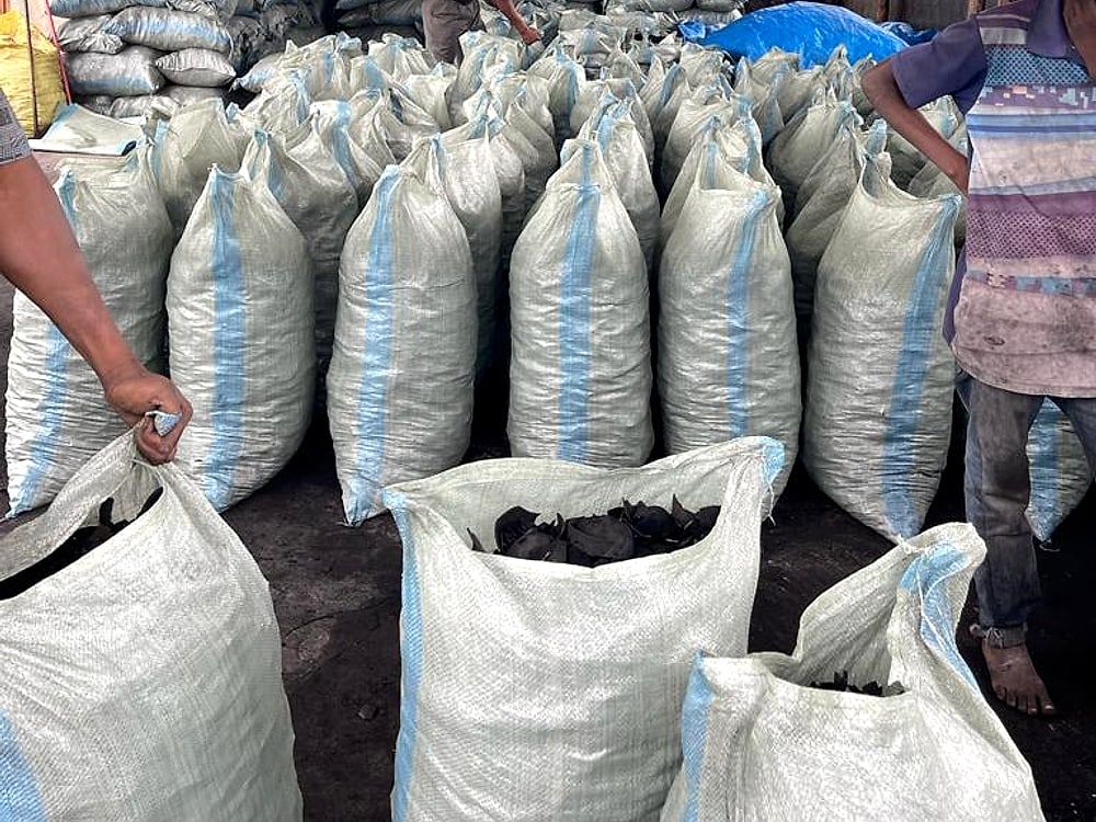 Workers filling woven sacks with coconut shell charcoal under an open shelter, filled sacks standing behind.