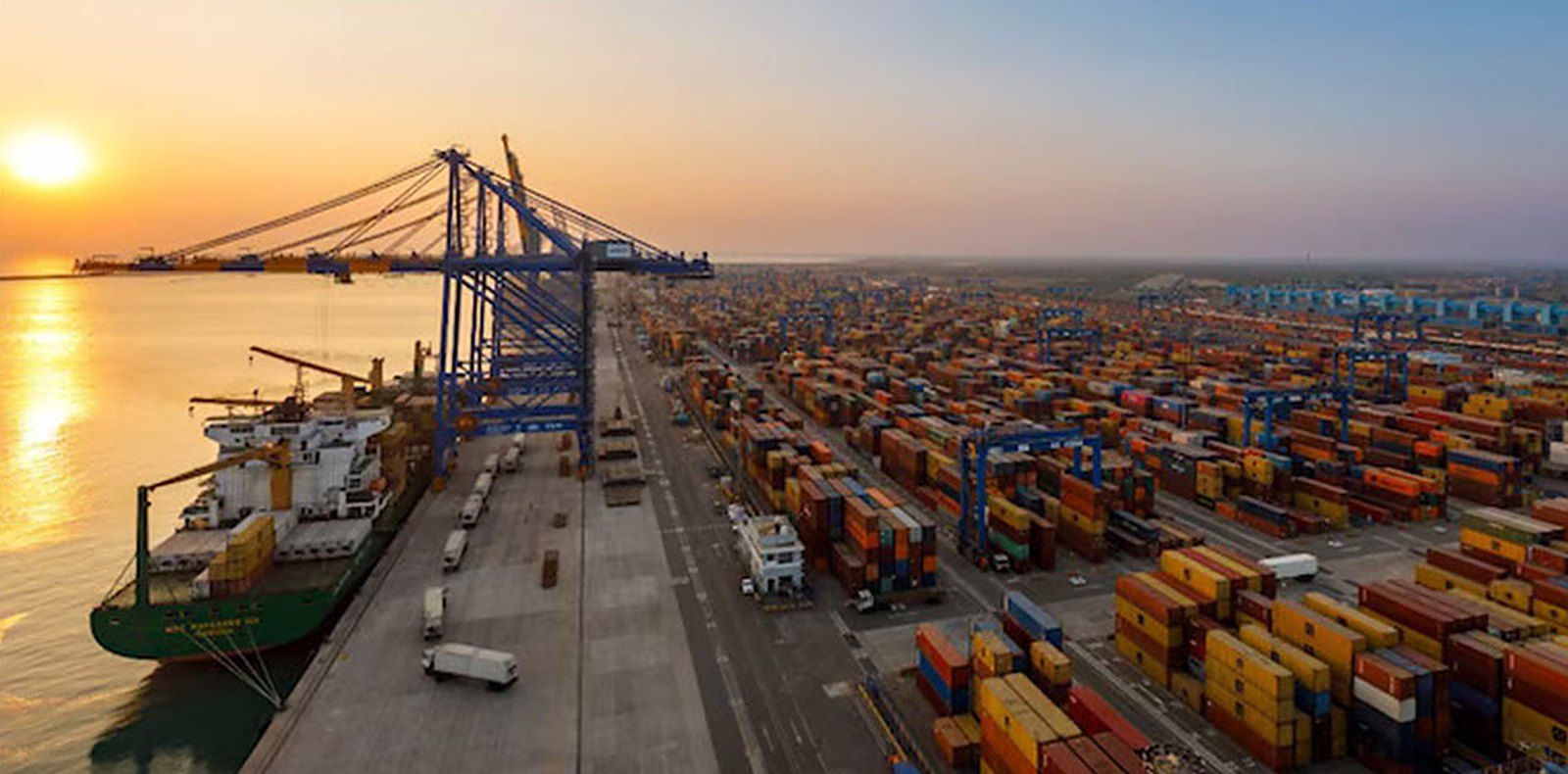 A container terminal at sunset: a ship alongside the quay under gantry cranes, with stacked containers running back to the horizon.